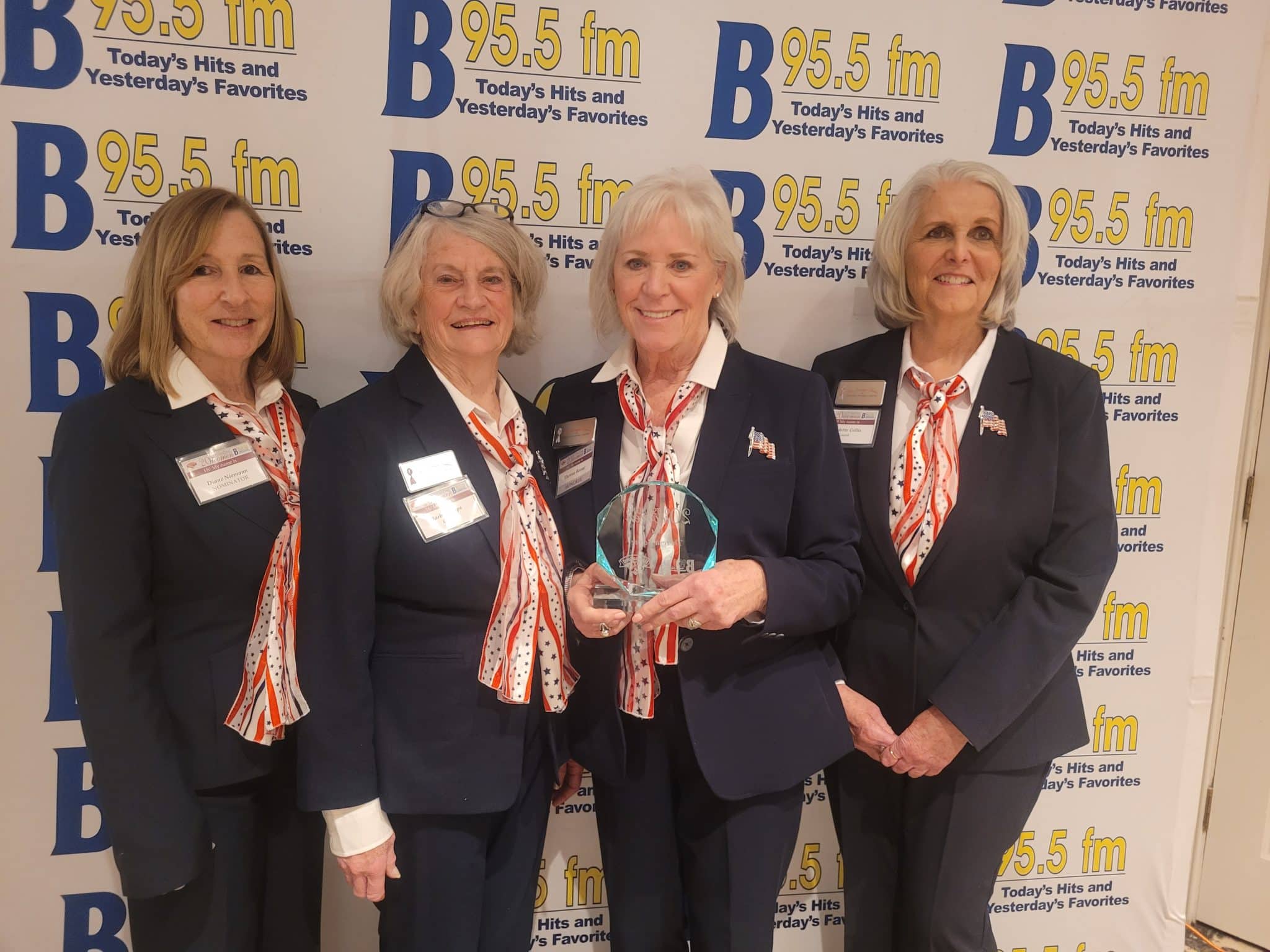 The Saratoga Ladies Four women in matching suits and red, white, and blue scarves pose together; one holds a glass award. A B 95.5 fm step-and-repeat backdrop is behind them.