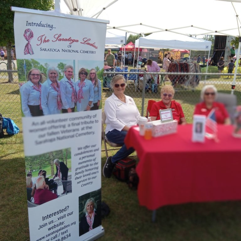 The Saratoga Ladies Three women sit at a table next to a Saratoga Ladies banner, supporting Saratoga National Cemetery, at an outdoor event with tents and people in the background.